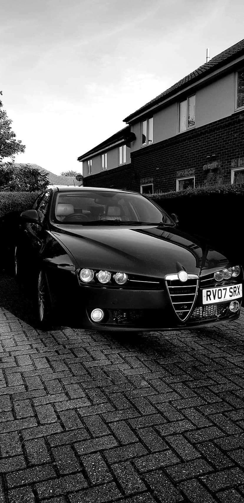 Black Alfa Romeo car parked on a cobblestone driveway with a modern house under an overcast sky in grayscale.