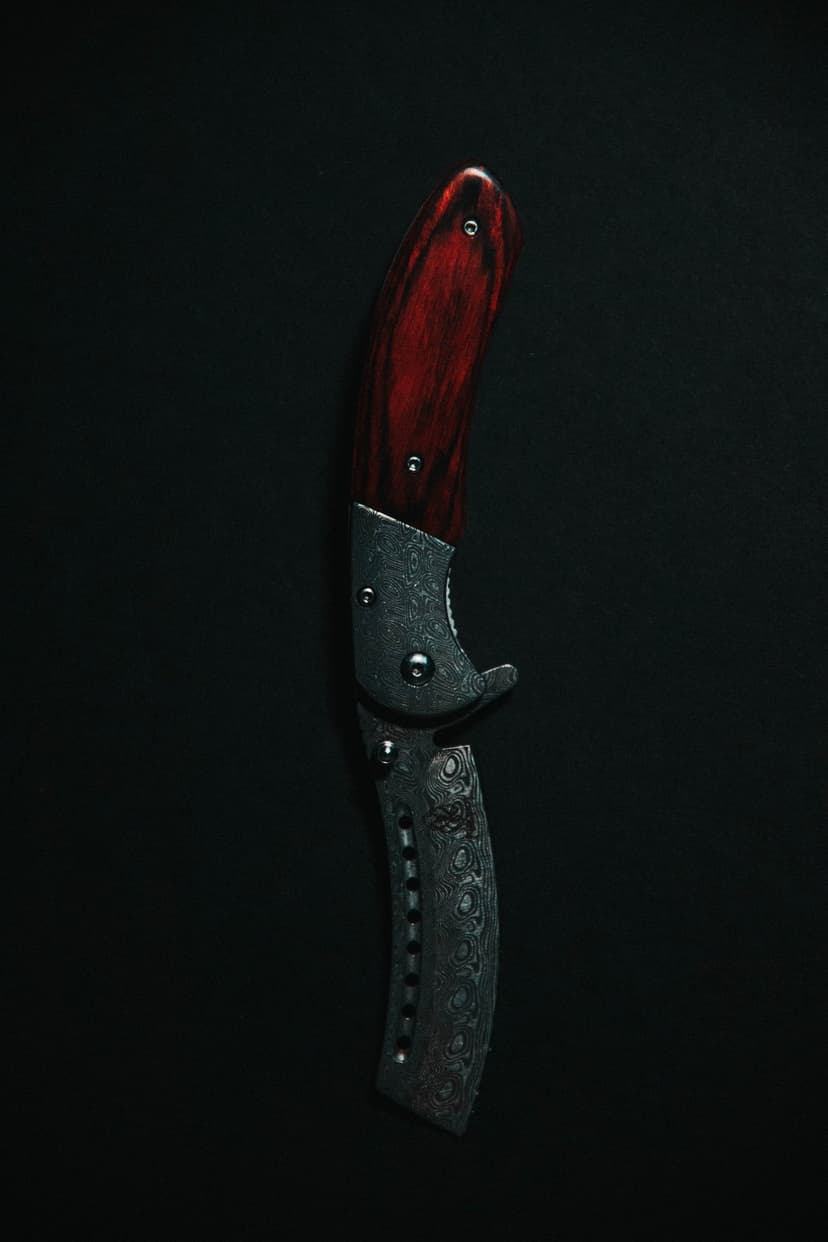 Damascus folding knife with a dark red wooden handle lies closed against a solid black, minimalist background.