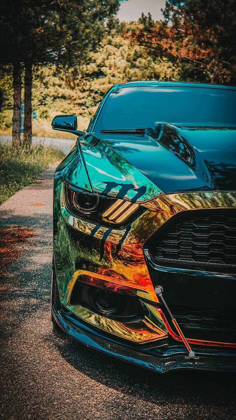 Ford Mustang with an iridescent chroma wrap and red tow straps parked on an asphalt path near green trees.