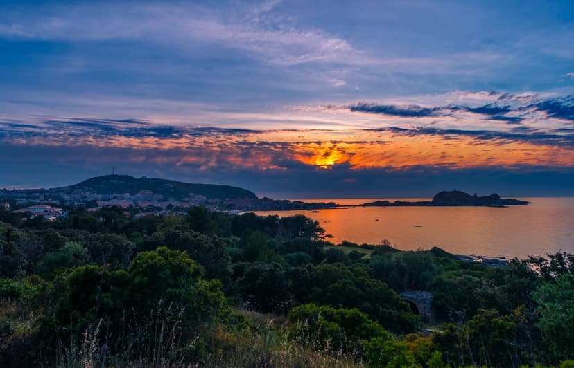 Coastal town silhouettes rest on a hillside above the Mediterranean Sea under a vibrant orange and purple sunset.