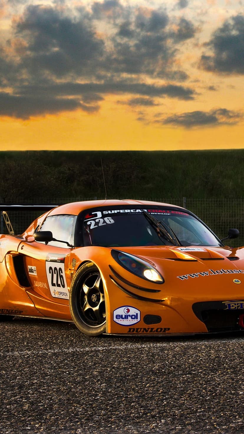 Orange Lotus race car 226 rests on a gravel circuit beneath a glowing sunset sky with dramatic cloud layers.