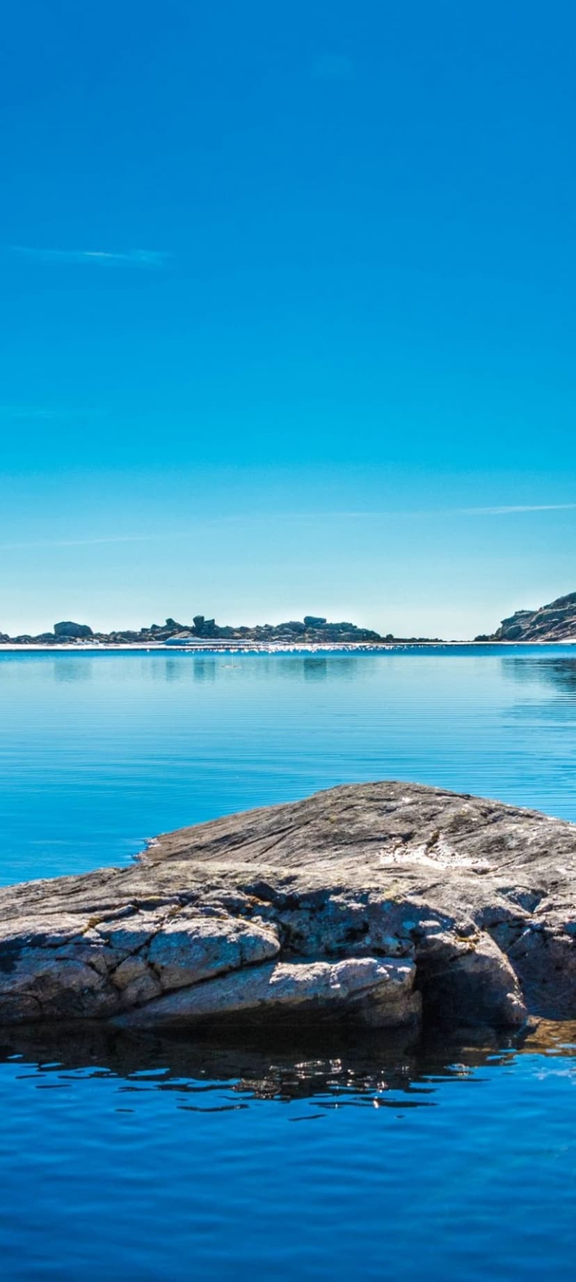 Weathered grey rocks sit in shallow blue water under a clear sky with a distant island chain on the horizon.