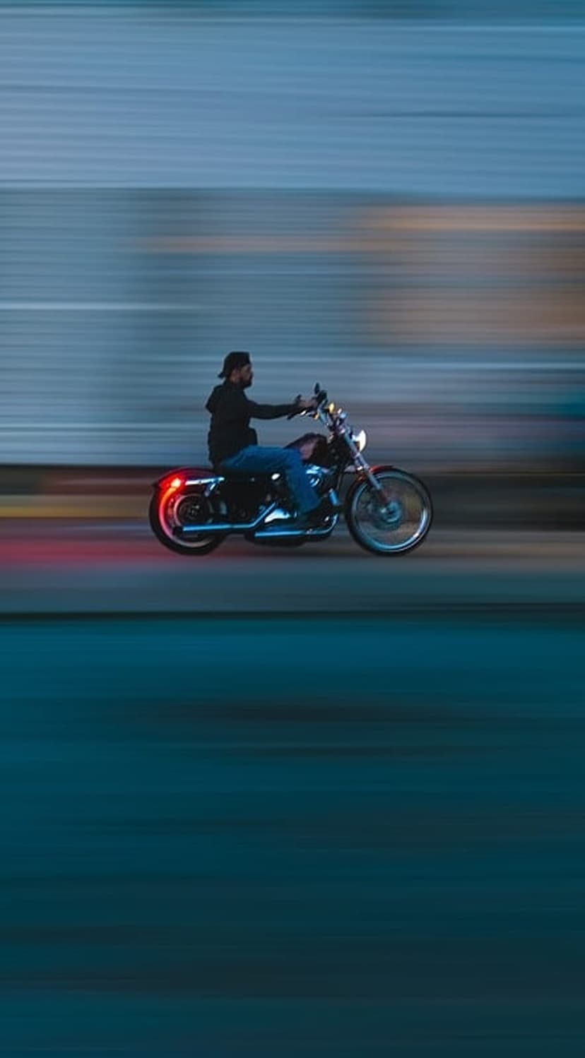 Motorcyclist in a black jacket speeds down a dark street with glowing red taillights and motion blur effects.