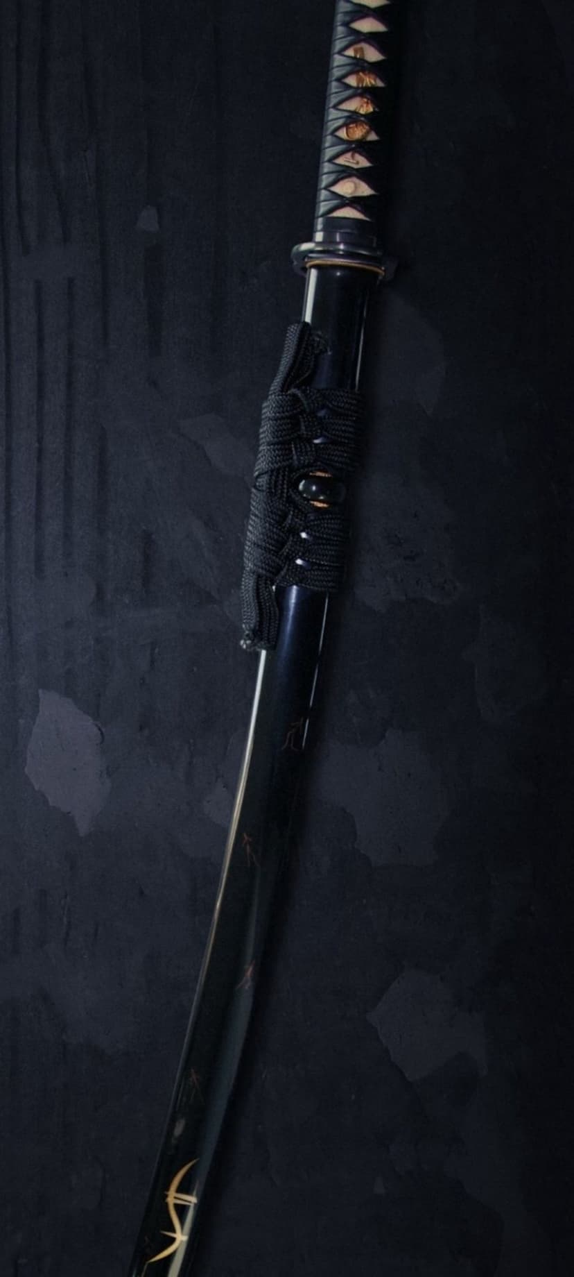 Samurai katana resting vertically against a dark textured wall with gold hilt accents and a black corded handle.