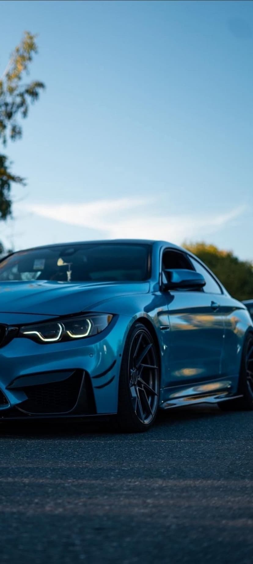 Blue BMW M4 Coupe with black wheels parked on a textured asphalt road under a blue sky and green trees.