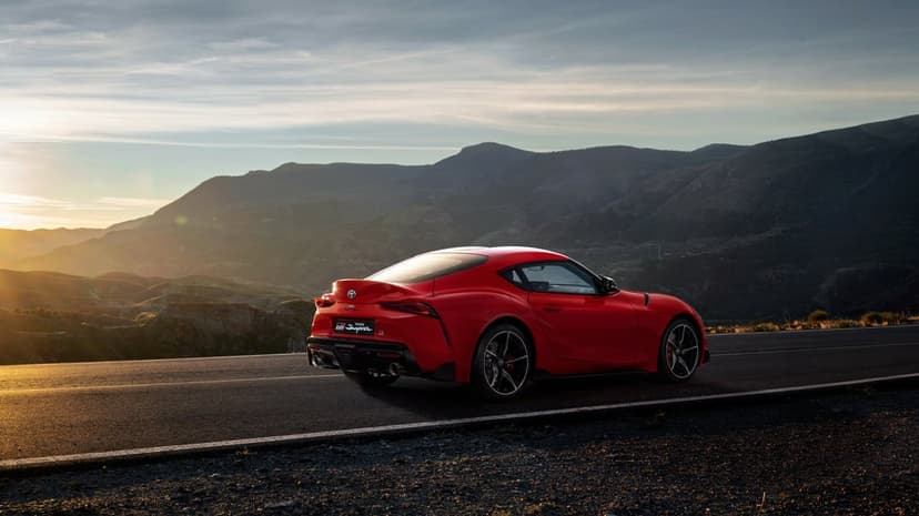 Red Toyota GR Supra on a mountain road at sunset with golden light and distant peaks in landscape.
