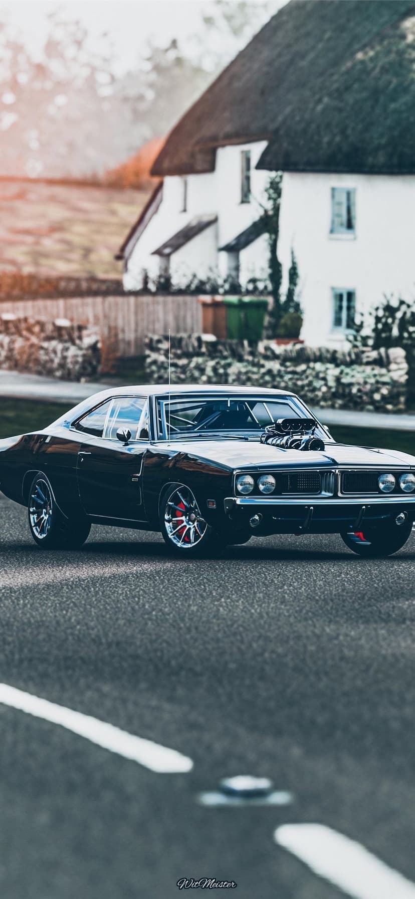 Black classic muscle car with a huge supercharger parked on a road in front of a white thatched roof cottage.