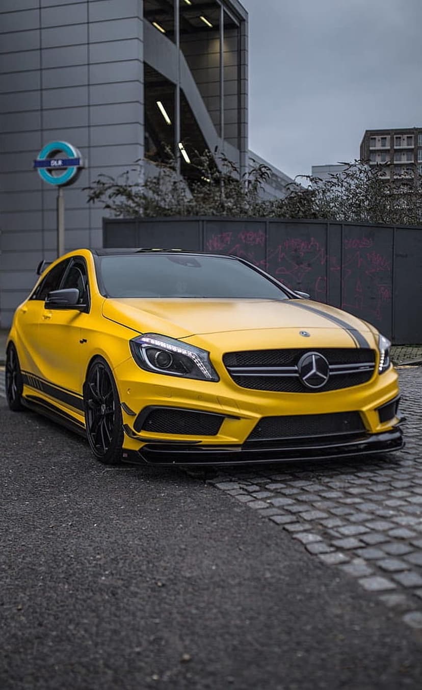 Yellow Mercedes-Benz AMG hatchback with black stripes parked on a cobblestone street near a London subway sign.