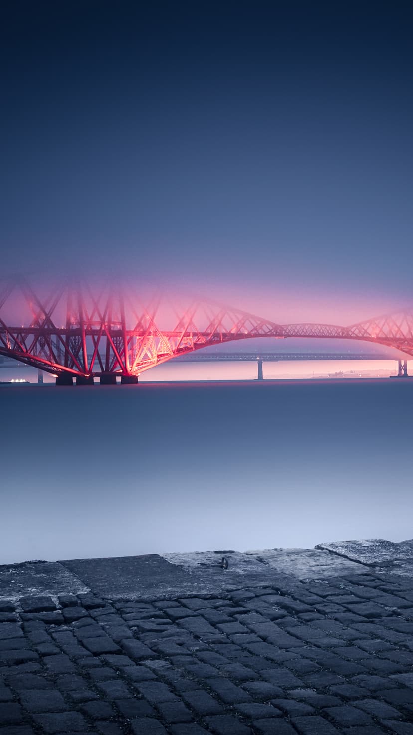 Forth Bridge glowing red under a twilight sky reflects in the dark Scottish estuary waters at dusk.
