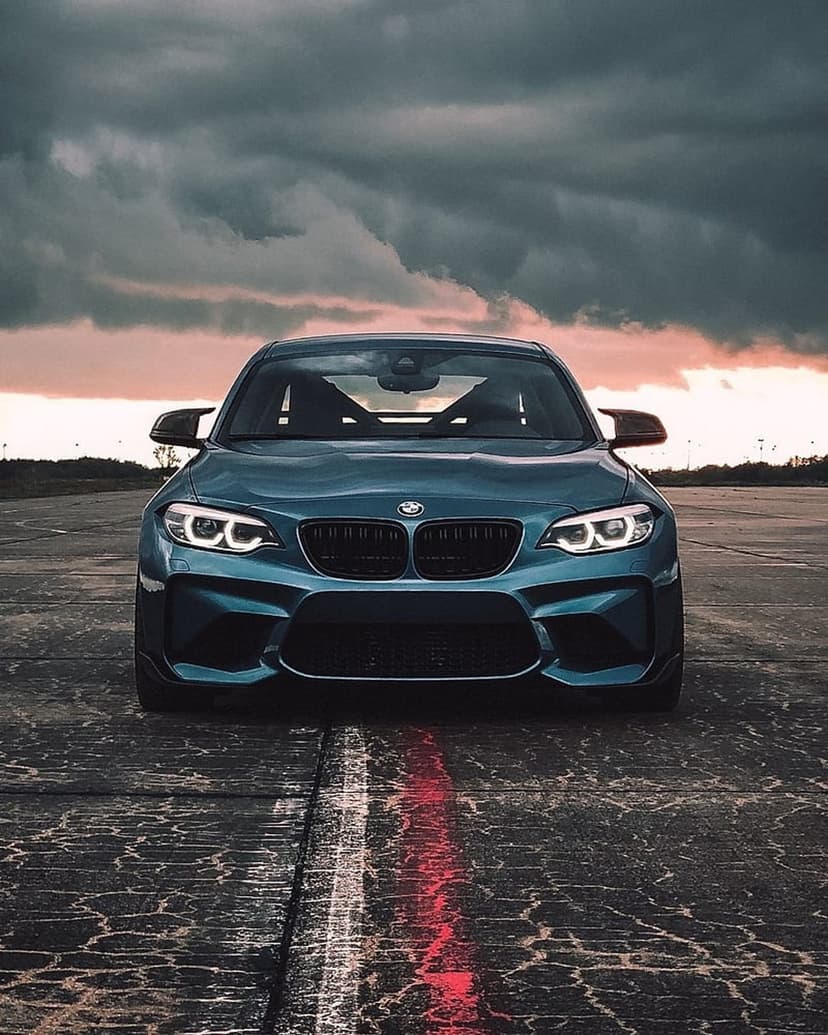 Blue BMW M2 parked on a striped asphalt road under a cloudy sunset sky, showcasing the front grille and headlights.