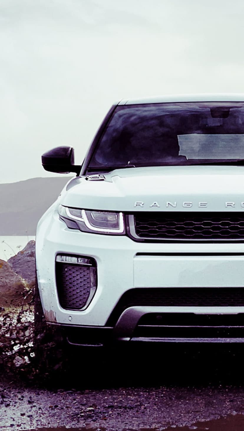 White Range Rover Evoque SUV kicking up mud and water on a rocky path under a cloudy sky during an off-road drive.
