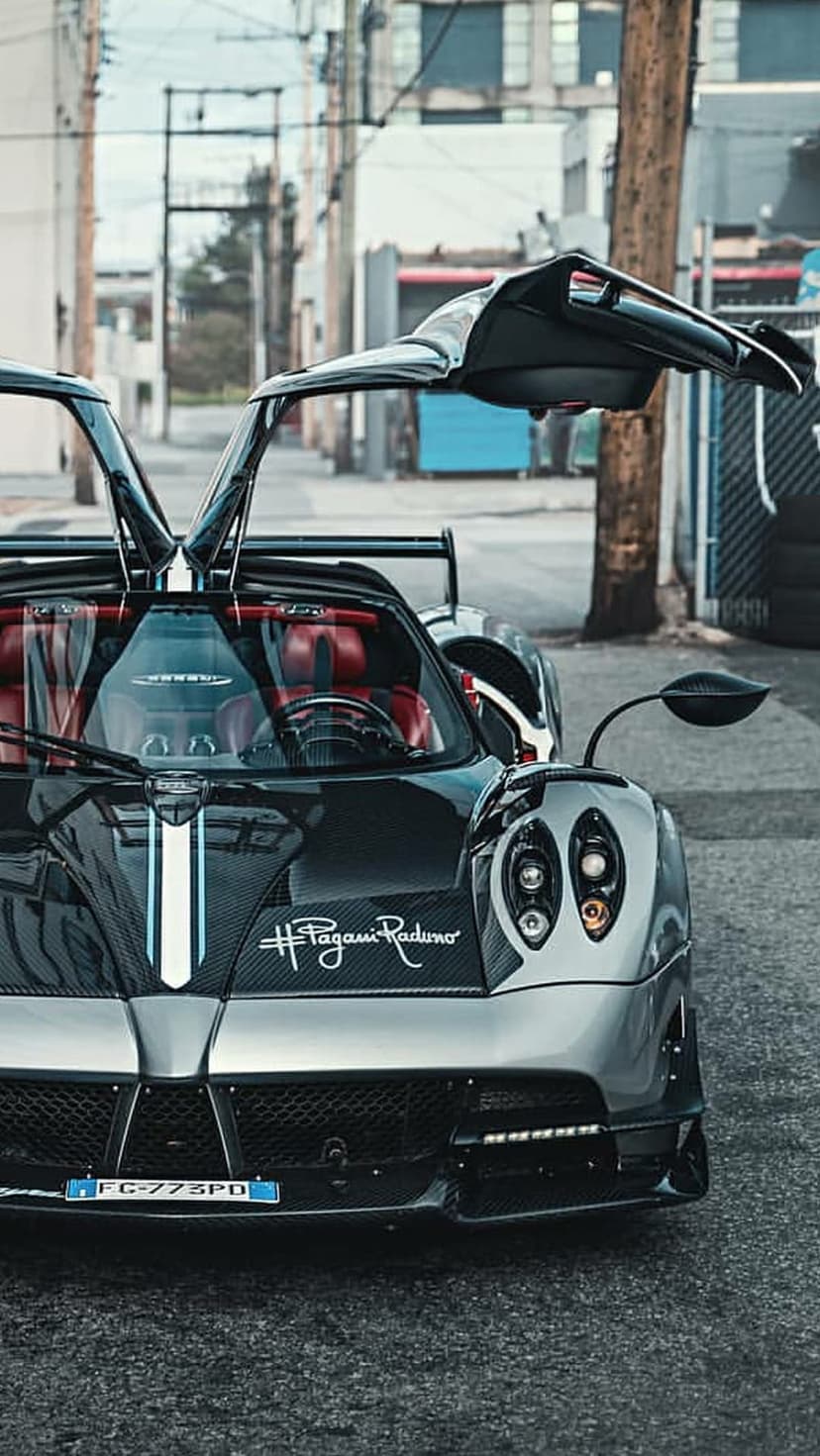 Pagani Huayra BC Roadster with open gullwing doors, red interior, and white racing stripe in a dark urban alley.