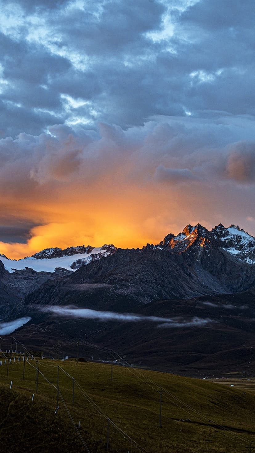 Snow-capped mountain peaks glow with orange sunset light beneath dark heavy clouds and green alpine slopes.