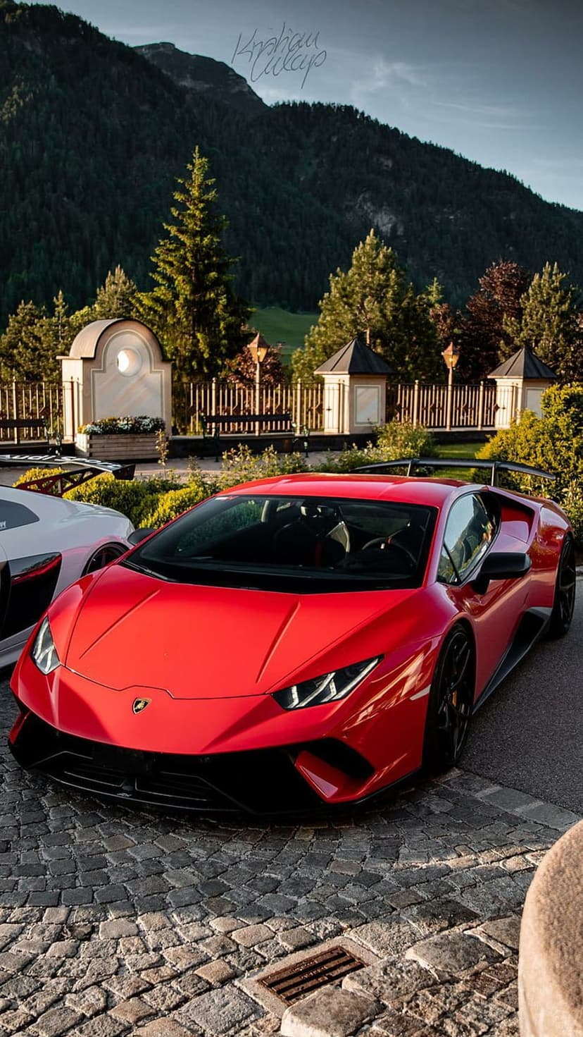 Red Lamborghini Huracan on a cobblestone path with lush green mountains and misty trees in the background.