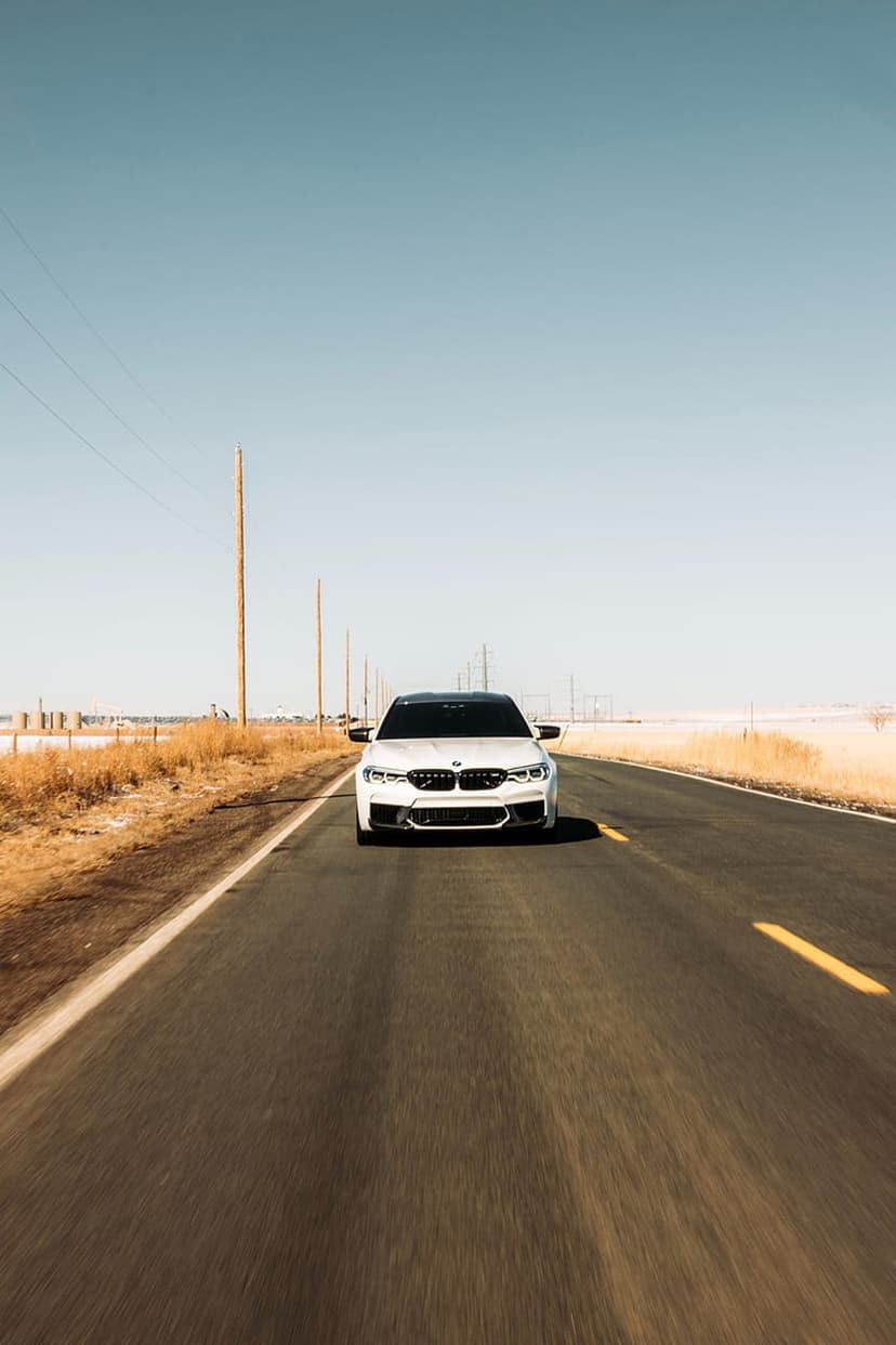 White BMW M5 sedan centered on a desert highway under a blue sky with dry grass lining the roadside.