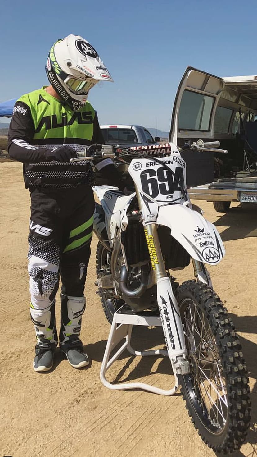 Motocross racer in full gear stands by a white dirt bike with number 694 on a sunny outdoor dirt track.