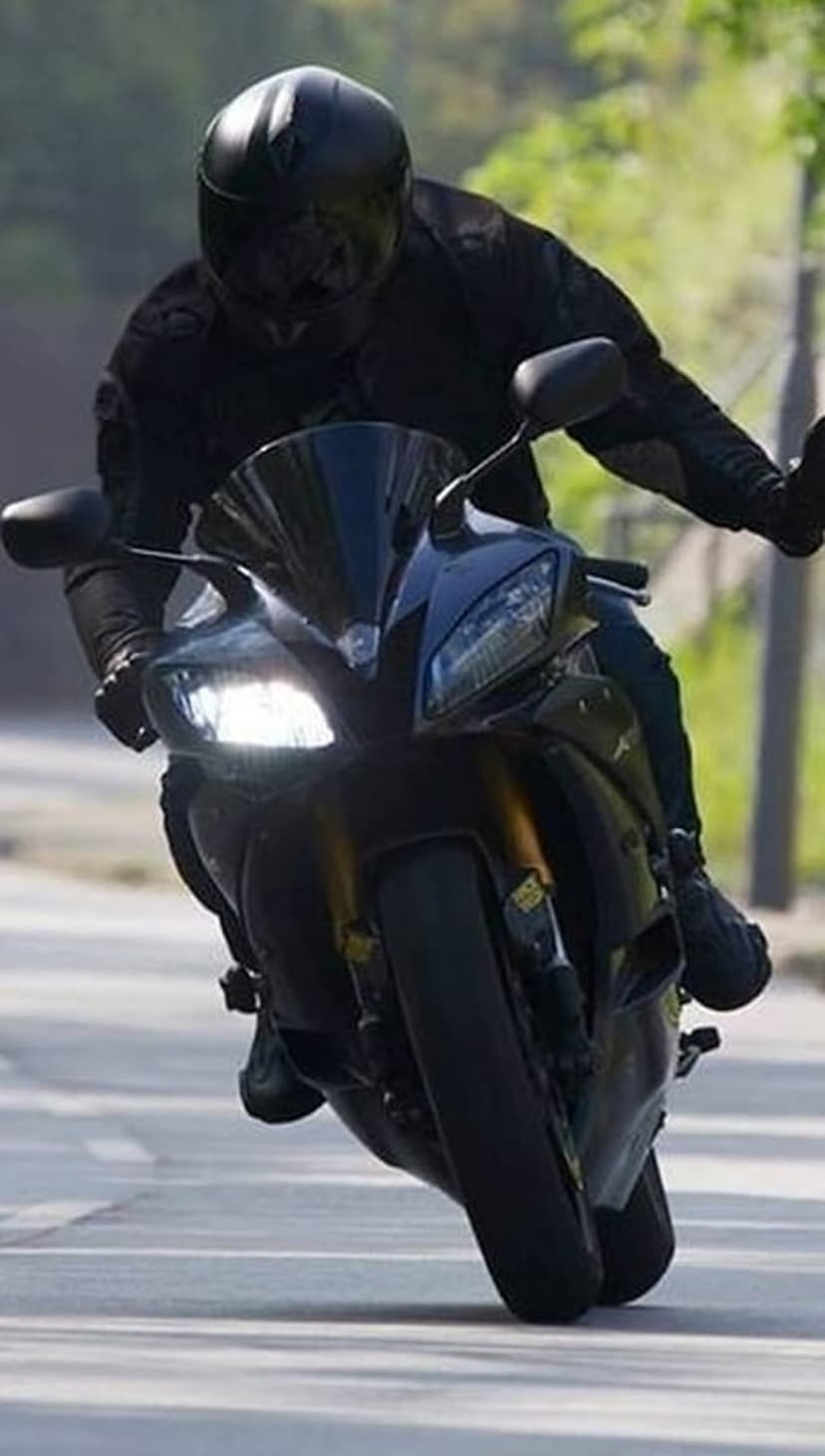 Motorcyclist in black leather gear leans low into a turn on a paved road with glowing motorcycle headlights.