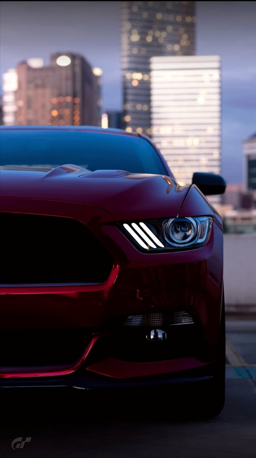 Red Mustang LED headlights glow at a low angle against a blurred background of illuminated city skyscrapers.
