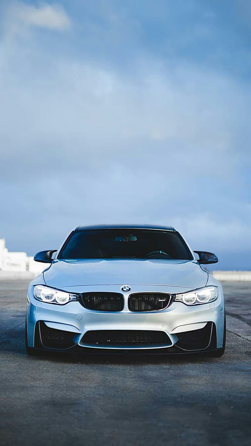 Silver BMW M4 with illuminated headlights parked on pavement under a cloudy sky for a phone background.
