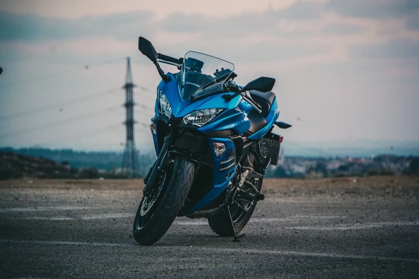 Blue Kawasaki sportbike parked on gravel with a tall industrial tower and city skyline under a cloudy sky.