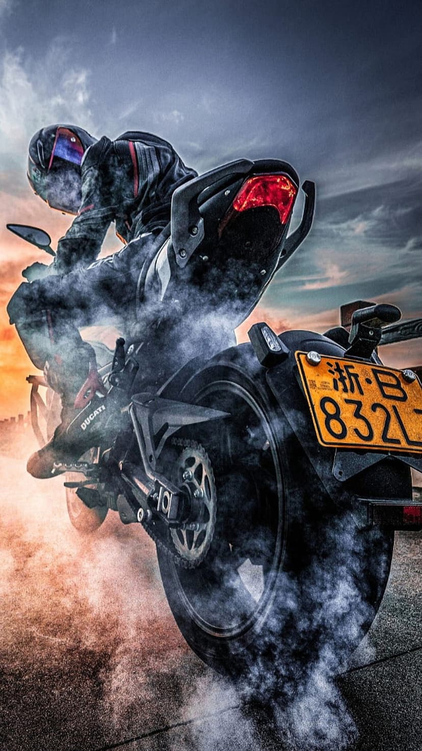 Motorcyclist in racing gear performs a burnout with white smoke against a vibrant orange and purple sunset sky.