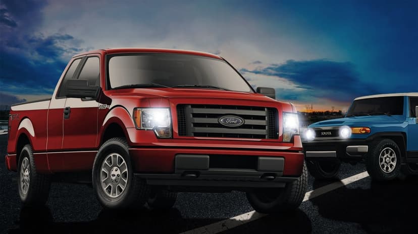 Red Ford F-150 and blue Toyota FJ Cruiser on a road at dusk with headlights shining under a cloudy sky.