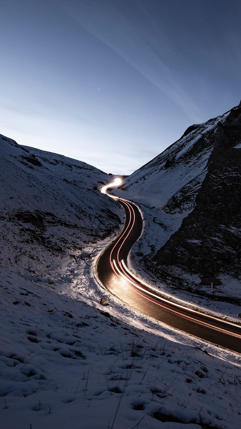 A winding mountain road with orange light trails cuts through snow-covered peaks under a dark blue twilight sky.