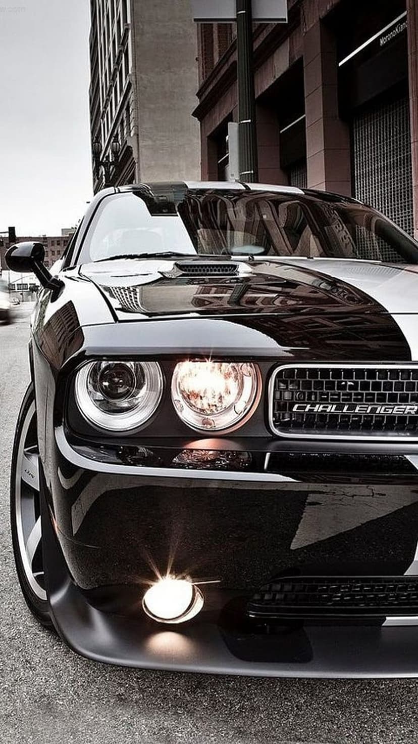 Black Dodge Challenger with a striped hood and circular headlights parked on a reflective city street at dusk.