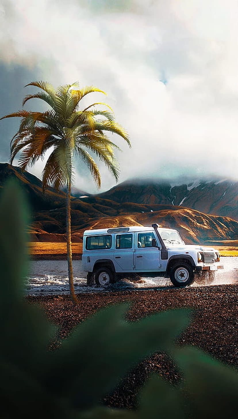 White Land Rover Defender driving through shallow water near palm trees with snowy mountains in the background.
