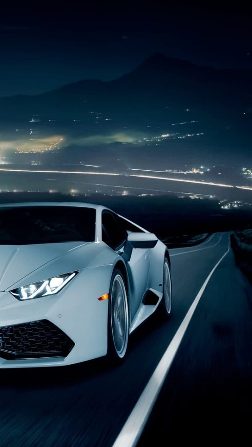 White Lamborghini racing on a winding mountain road at night with distant city lights twinkling below.