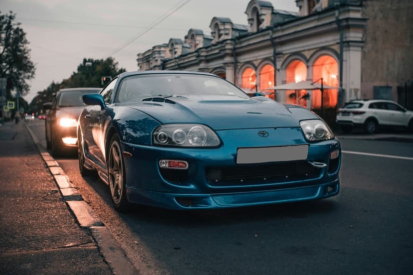 Blue Toyota Supra parked on an asphalt city street at dusk with a vintage building and arched windows behind it.