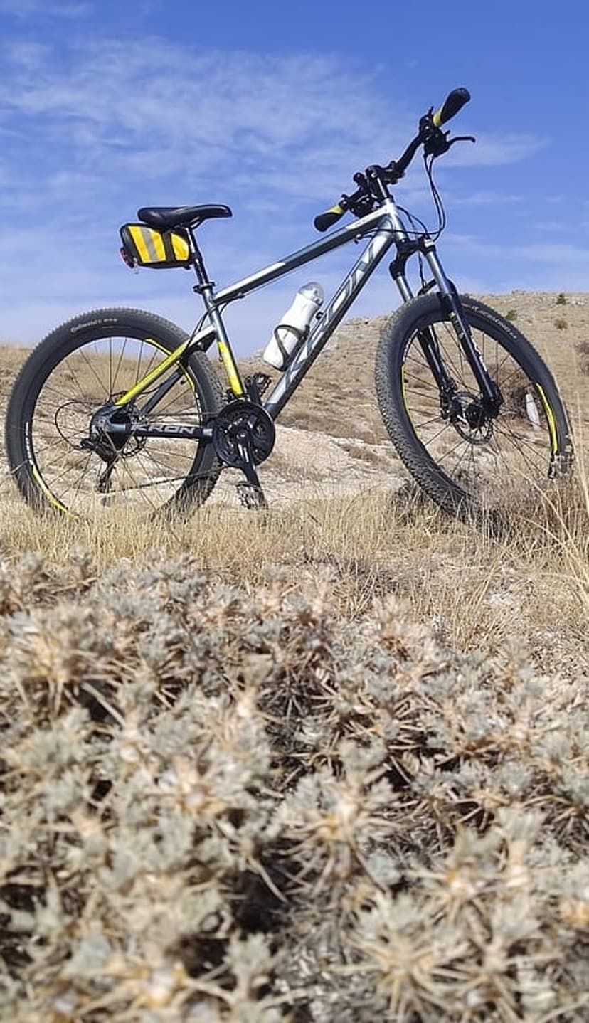 Silver and yellow mountain bike parked on dry grass with rolling arid hills under a clear blue sky background.