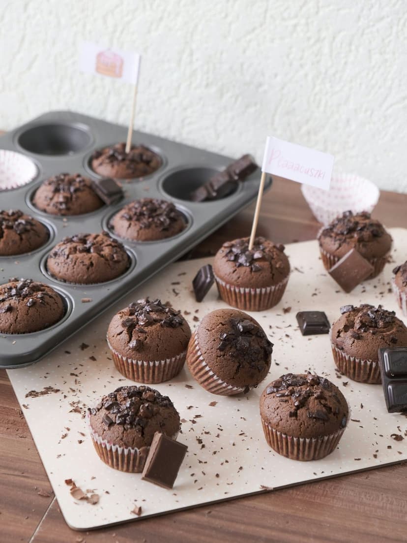 Chocolate muffins topped with large cocoa chunks and small paper flags rest on a rustic parchment surface.