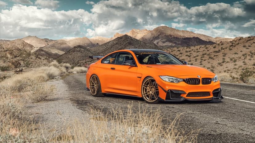 Orange BMW M4 with bronze wheels parked on a desert road with mountains and a cloudy sky in the background.