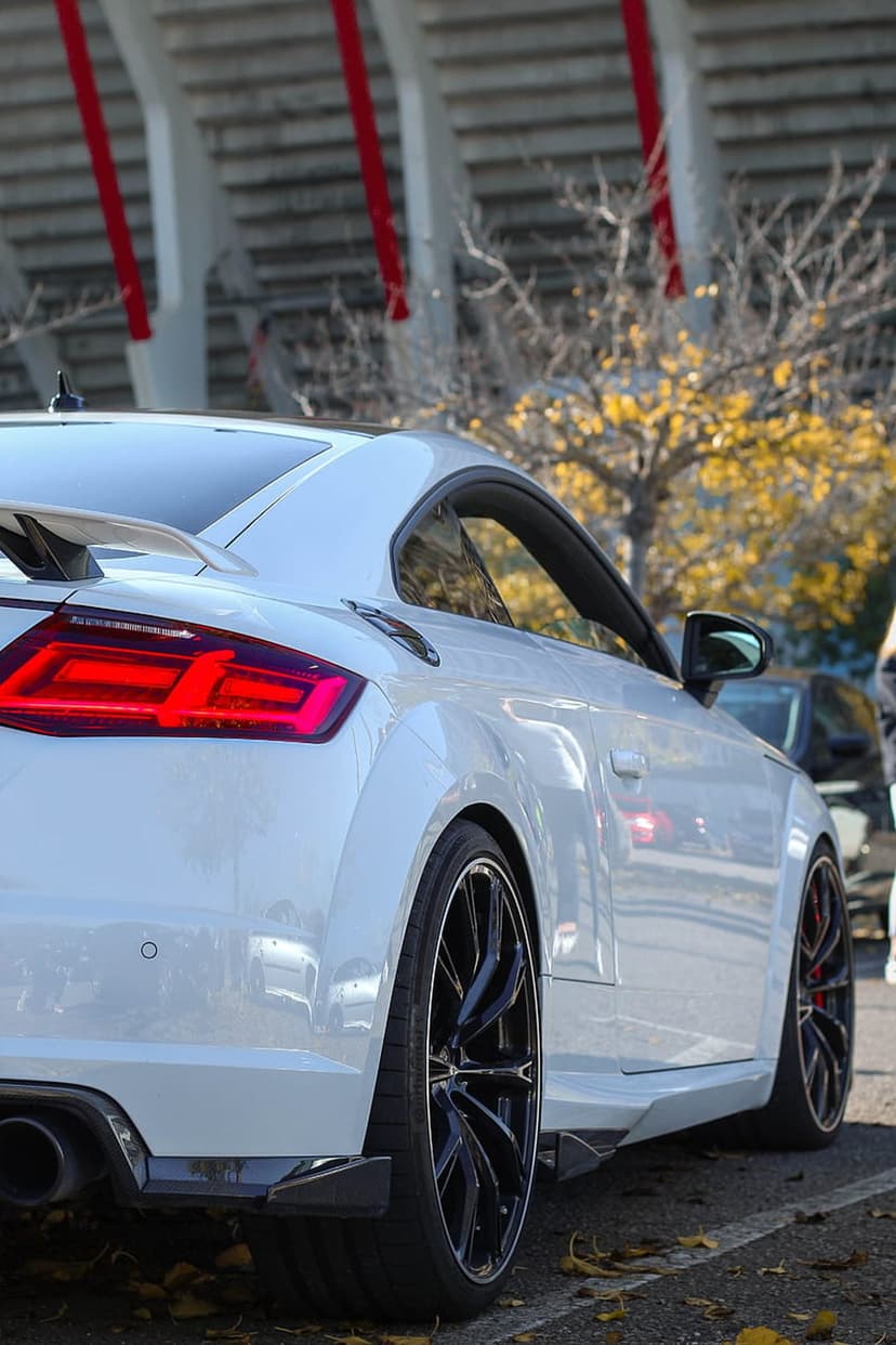 White Audi TT RS Coupe with glowing red taillights and black wheels parked near stadium seating and fall trees.