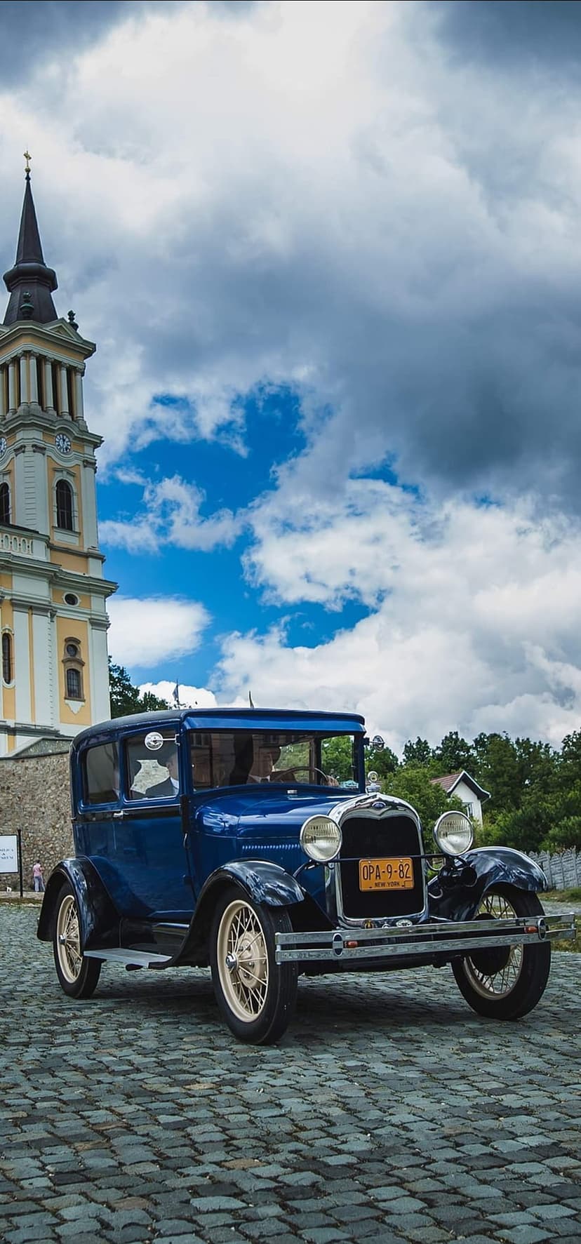 Blue Ford Model A sedan parked on a cobblestone street with a tall stone church tower under a stormy sky.