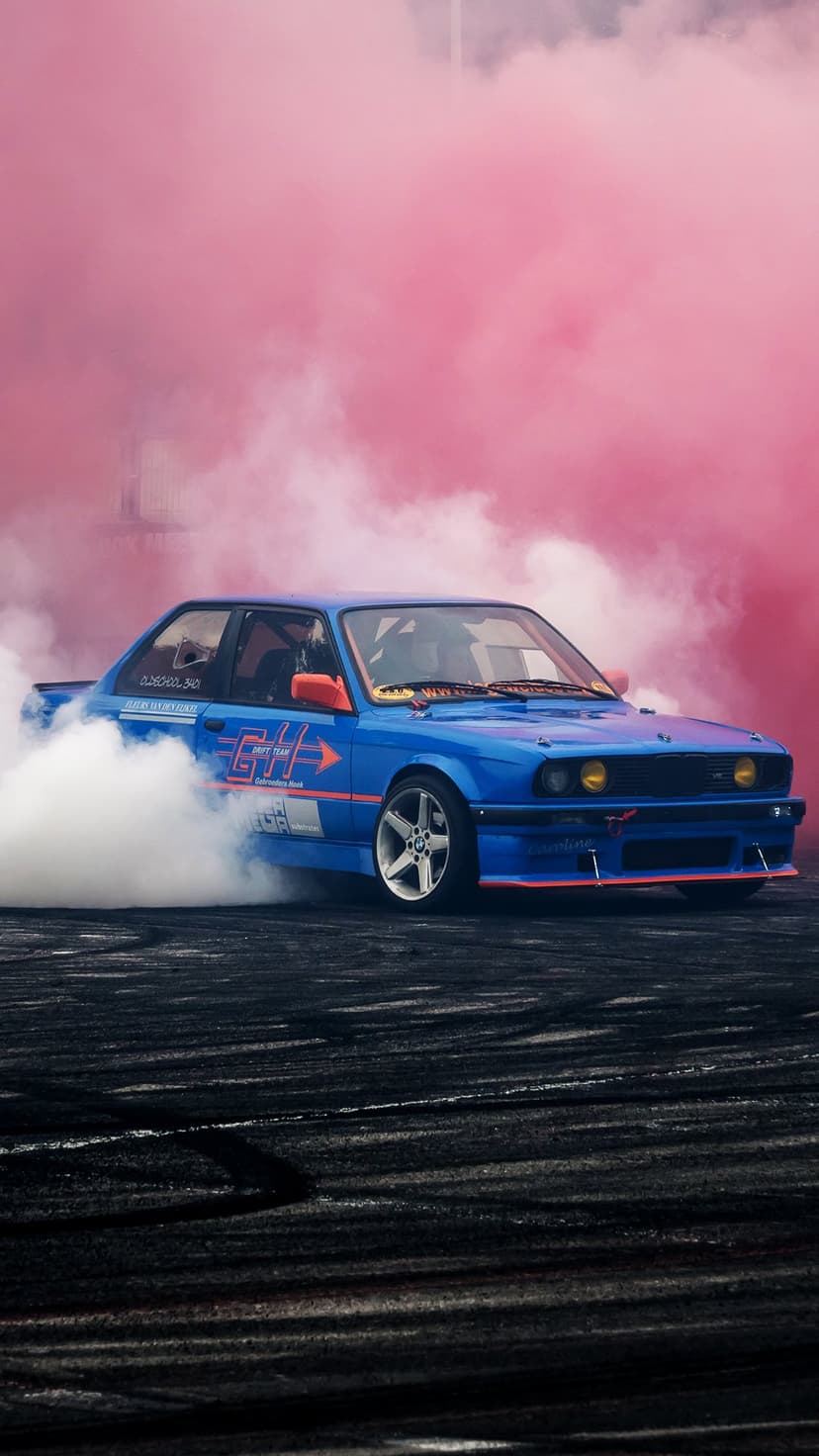 Blue drift car with orange accents skidding on dark asphalt while emitting thick white and pink tire smoke clouds.