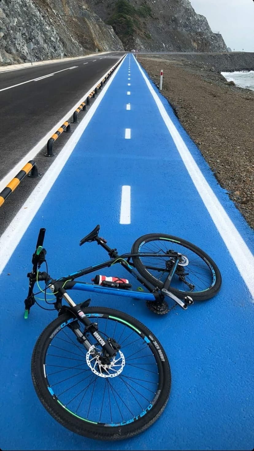 Blue cycling path with a bicycle lying down curves along a rugged mountain cliff and the turquoise ocean water.