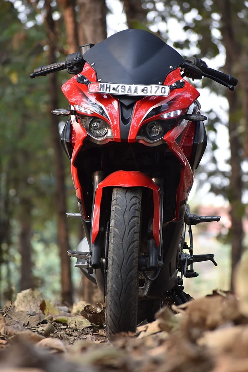 Red sports motorcycle with dual headlights parked on a bed of fallen leaves in a dense, sunlit forest grove.
