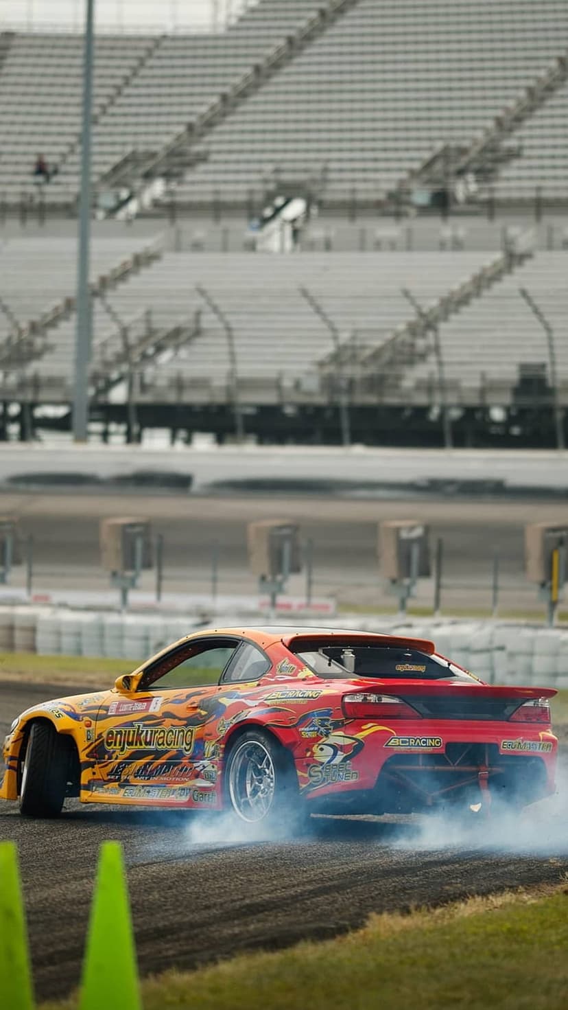 Orange drift car sliding on a race track with thick white smoke clouds behind tires and a stadium background.
