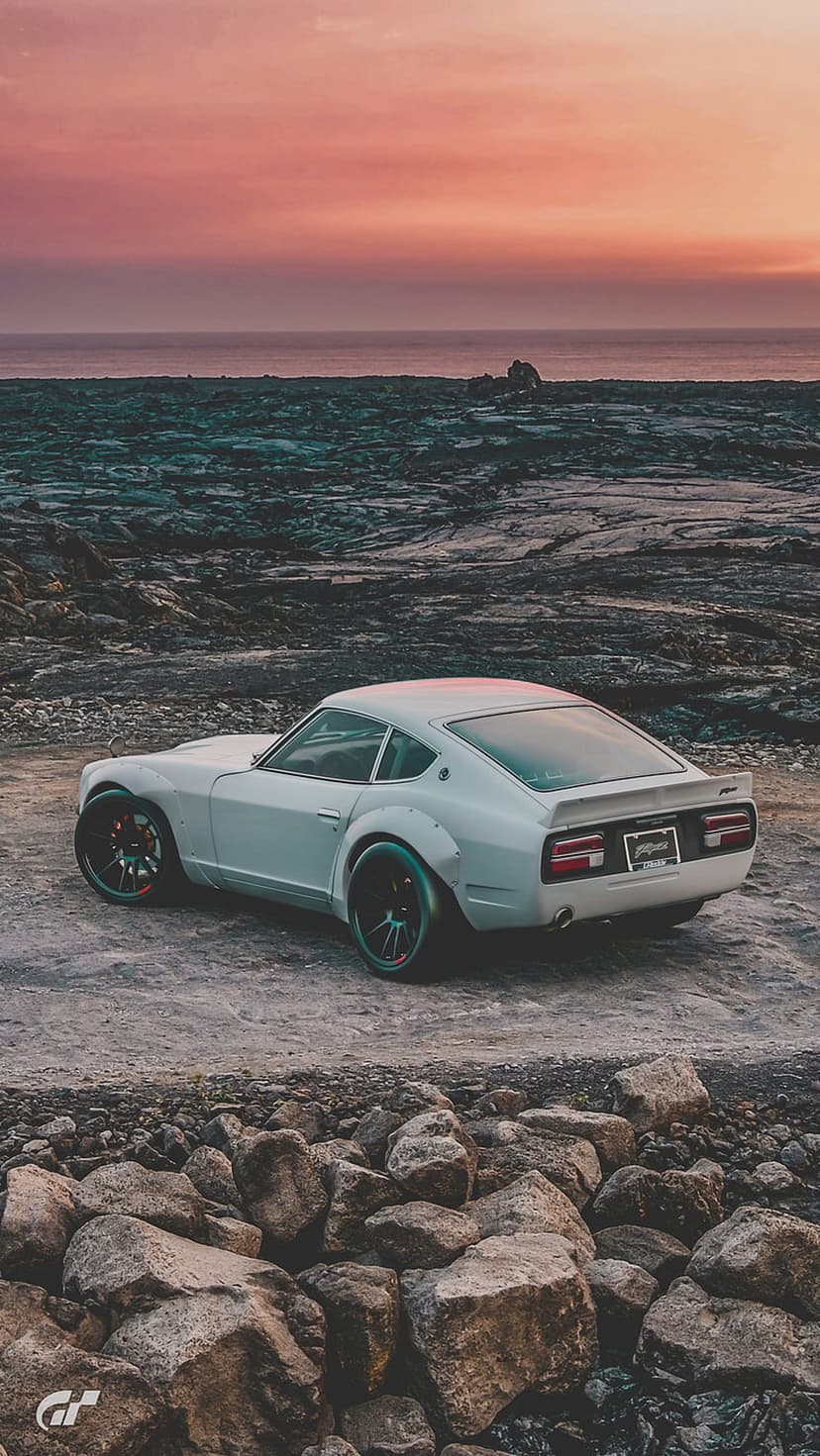 White Nissan Fairlady Z S30 with widebody fenders parked on jagged coastal rocks against a vibrant sunset sky.