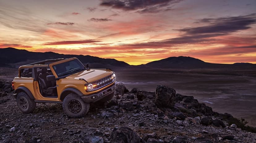 Orange off-road SUV parked on a rocky cliff under a dramatic sunset with purple and red clouds.