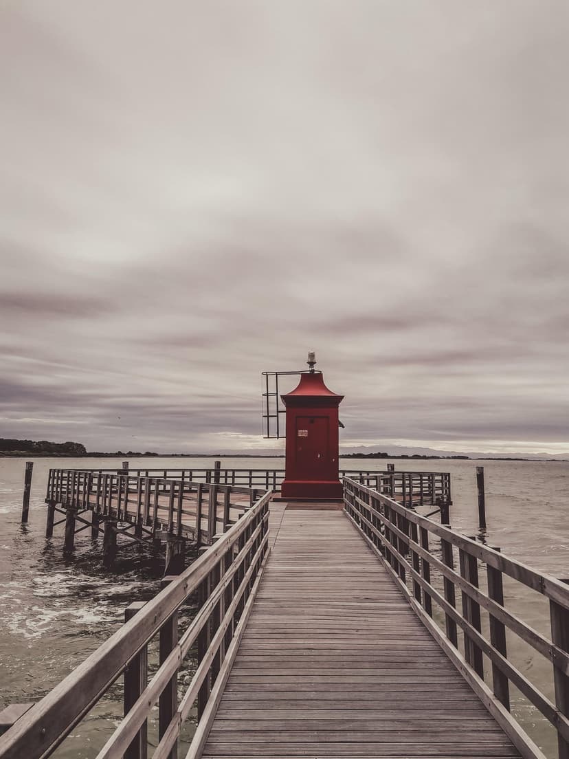 Red lighthouse at the end of a wooden pier under a cloudy sky above calm water in a vertical coastal scene.