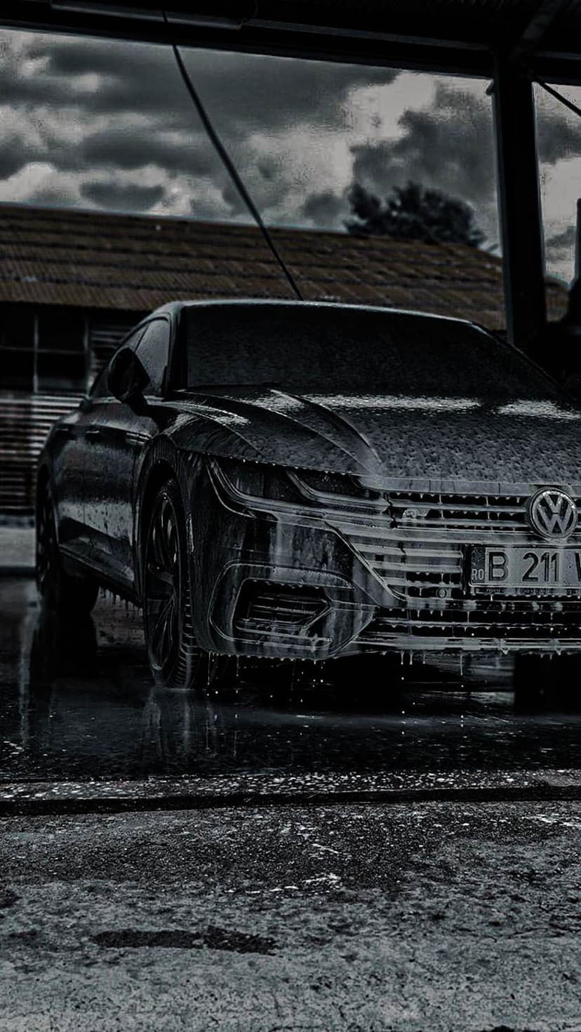 Black Volkswagen Arteon sedan covered in white soap foam on wet asphalt under a dark and moody overcast sky.