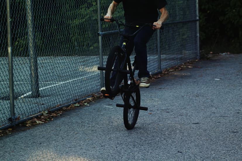 BMX rider in dark clothes balances a black bike on one wheel on a paved path next to a chain-link fence.