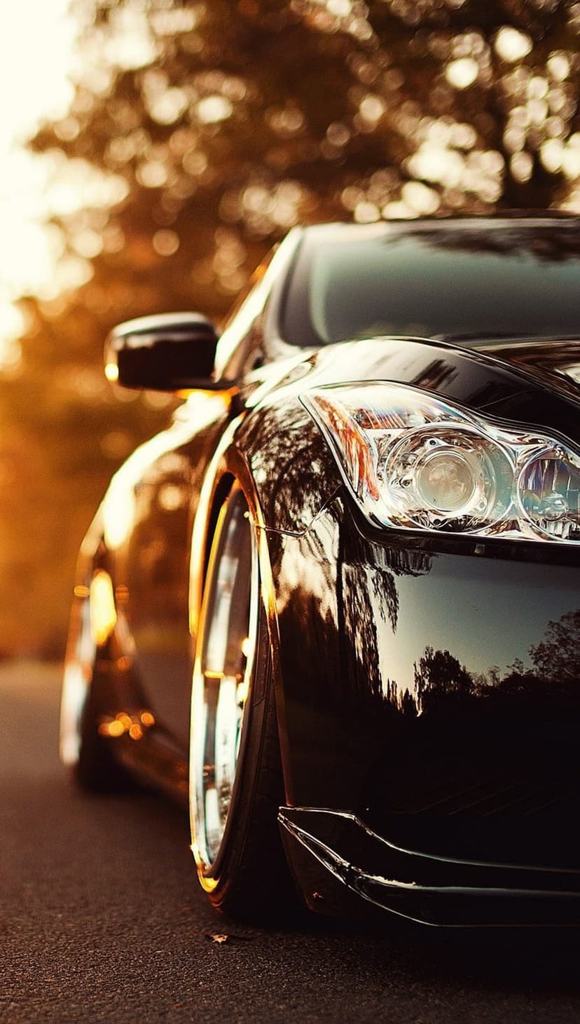 Black luxury car headlight and wheel rim reflect golden sunset light with warm bokeh in the background.