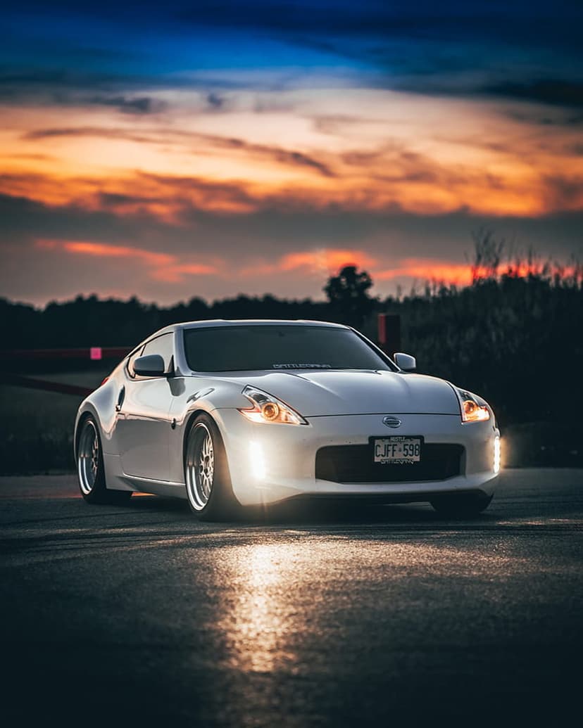 White Nissan 370Z sports car parked on a road at sunset with glowing headlights under a pink and orange sky.