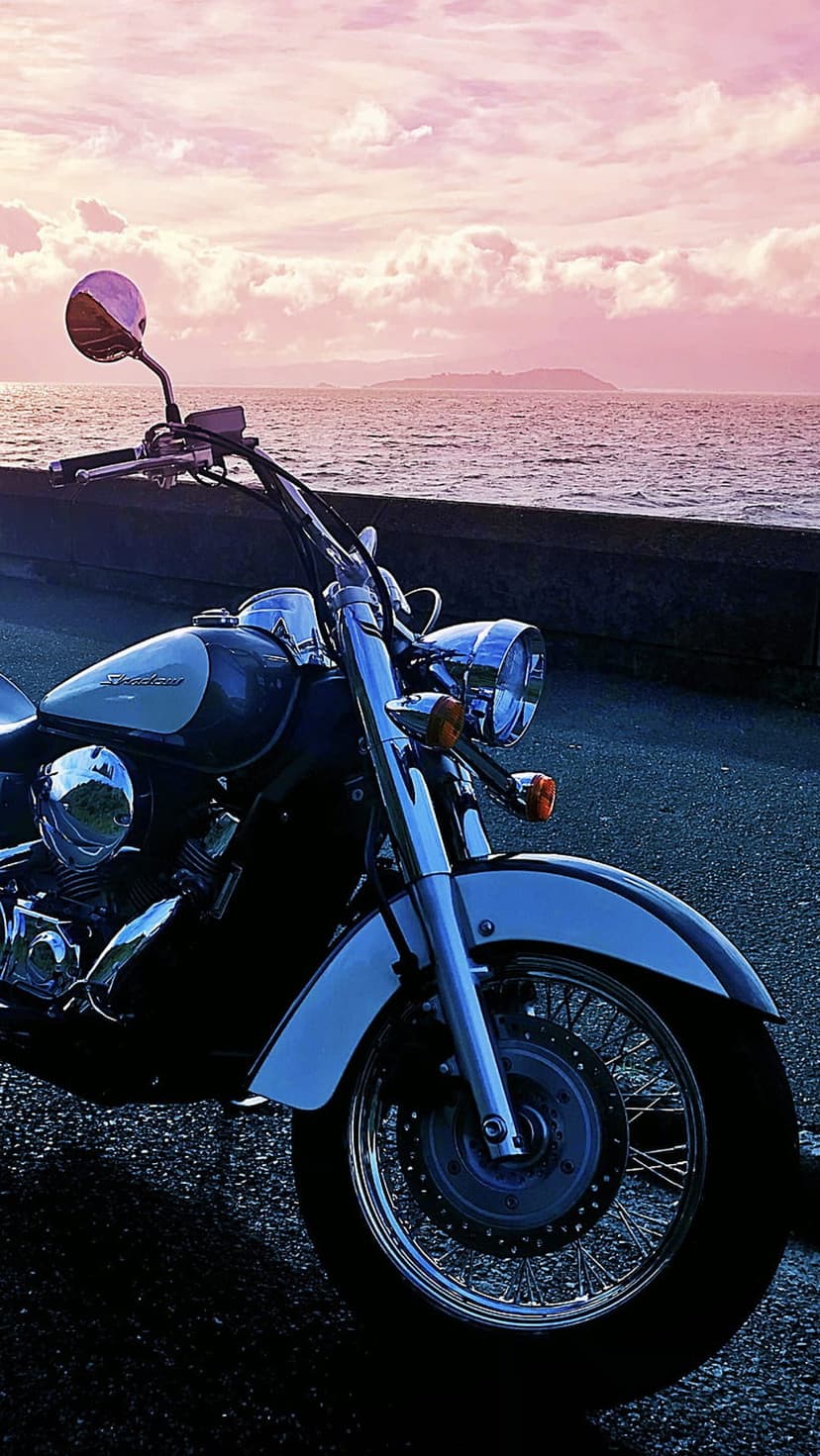Cruiser motorcycle with chrome accents parked on a coastline during a pink sunset with calm water reflections.