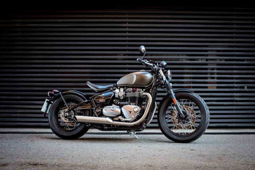 Profile of a grey Triumph Bonneville Bobber motorcycle against a dark slatted wall on an asphalt surface.