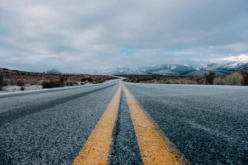 Asphalt road with double yellow lines leads toward jagged snow-covered mountains under a heavy cloudy gray sky.